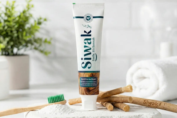 Tube de dentifrice naturel SiwakSmile et bâtons de miswak traditionnels disposés sur une pierre blanche à côté d'une brosse à dents en bambou et de poudre, illustrant une routine de blanchiment dentaire naturel dans une salle de bain lumineuse.