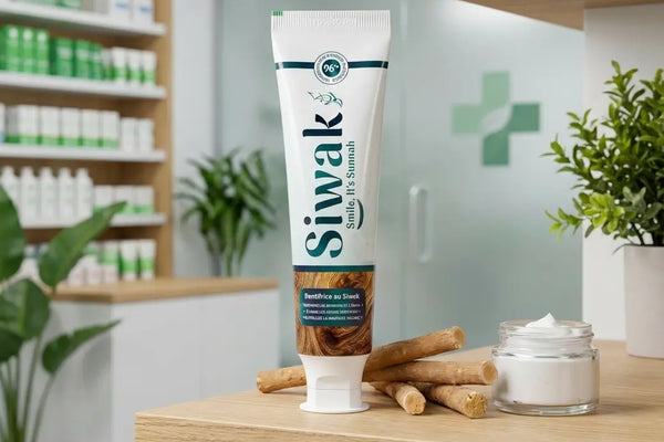 Tube de dentifrice naturel Siwak "Smile, it's Sunnah" posé sur une étagère en bois, entouré de bâtons de siwak bruts et d'un petit pot de pâte, dans un environnement de pharmacie avec une croix verte en arrière-plan.